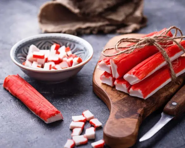 Paluszki krabowe na desce, w miseczce i obok. Czy mrożone paluszki krabowe trzeba gotować? Zazwyczaj nie, są już gotowe do spożycia.