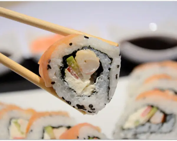 Chopsticks trzymają sushi z krewetkami, awokado i kremowym serkiem. W tle widać więcej roladek sushi.