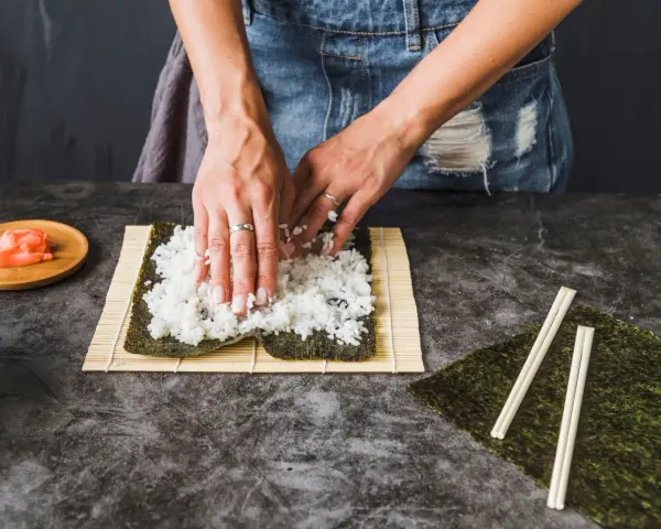 Dłonie rozprowadzają ryż na nori, przygotowując się do zwinięcia sushi. Obok leżą pałeczki i marynowany imbir, idealny dodatek do sushi.