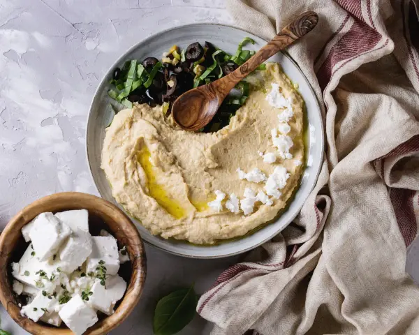 Kremowy hummus z ciecierzycy, posypany fetą i oliwkami, z drewnianą łyżką. Obok miseczka z kostkami sera feta.