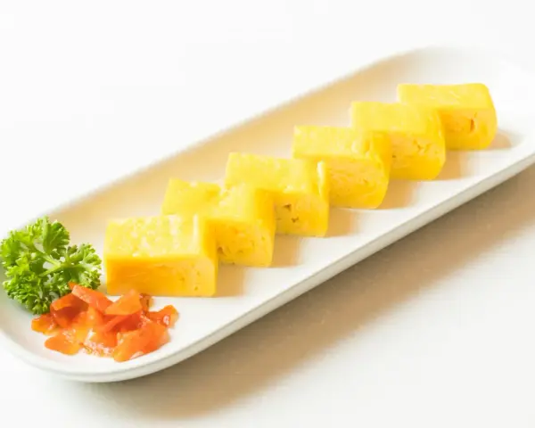 Sześć kawałków tamago, czyli japońskiego omletu, ułożonych na białej desce. Obok natka pietruszki i pomarańczowe dodatki.