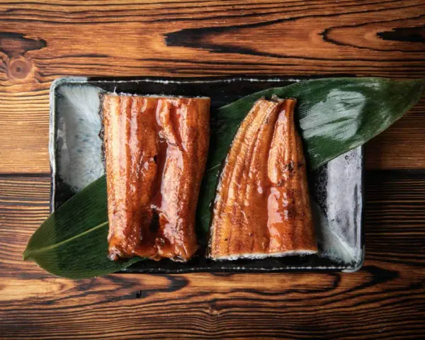 Dwa kawałki błyszczącego, glazurowanego sushi z węgorzem na liściu, podane na czarnej tacce.
