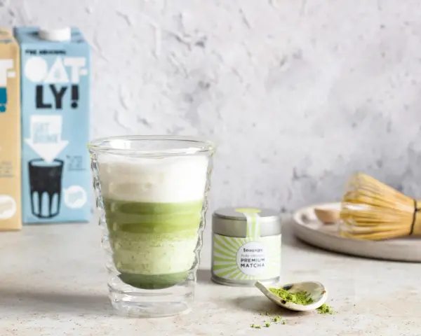 Orzeźwiający iced matcha latte z pianką, obok puszka matcha i bambusowa miotełka. Idealny napój na ciepłe dni.