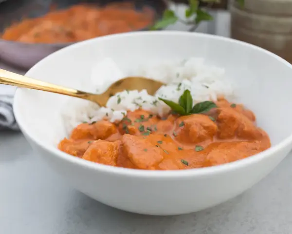 Aksamitny butter chicken masala z ryżem, udekorowany miętą. Złota łyżka zanurzona w daniu.