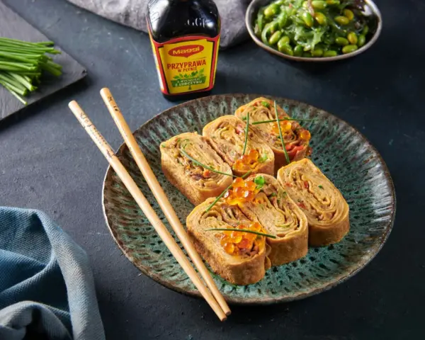 Roladki z omletu do sushi, ozdobione ikrą łososia i szczypiorkiem, obok pałeczki i sosu Maggi.