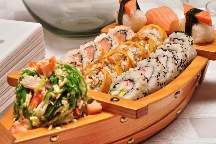 Drewniana łódka pełna sushi, dania z kraju, z którego pochodzi sushi, czyli Japonii. Obok stos białych talerzy.
