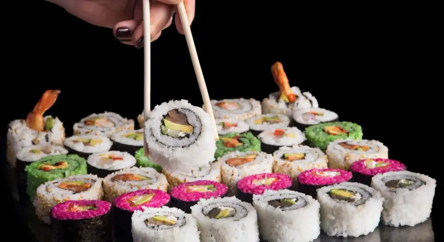 Dłoń z pałeczkami podnosi sushi. Różnorodne rolki, w tym zielone i różowe, czekają na spróbowanie. Nauka jak jeść pałeczkami to klucz do tej uczty.