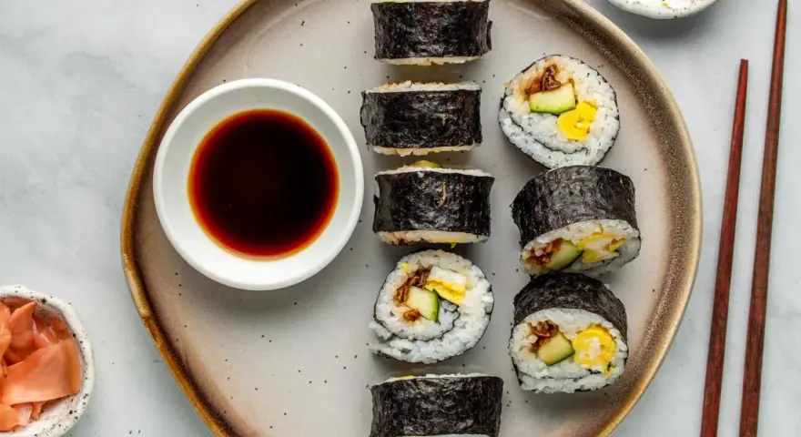 Na talerzu ułożone są sushi: kilka mniejszych hosomak i większe futomaki, obok miseczka sosu sojowego i marynowanego imbiru.
