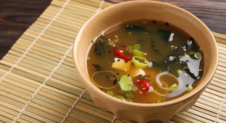 Aksamitna zupa miso z tofu, wodorostami, szczypiorkiem i papryczką chili, podana w ceramicznej miseczce na bambusowej macie.
