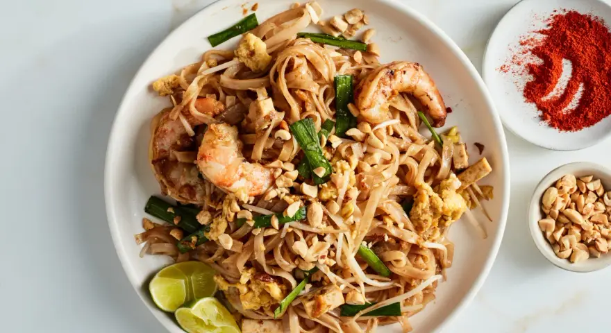 Talerz z Pad Thai z krewetkami, orzeszkami ziemnymi, jajkiem i limonką, obok miseczki z orzeszkami i papryką.