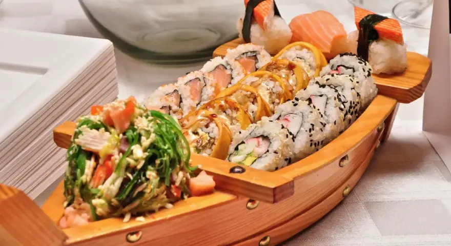 Drewniana łódka pełna sushi, dania z kraju, z którego pochodzi sushi, czyli Japonii. Obok stos białych talerzy.