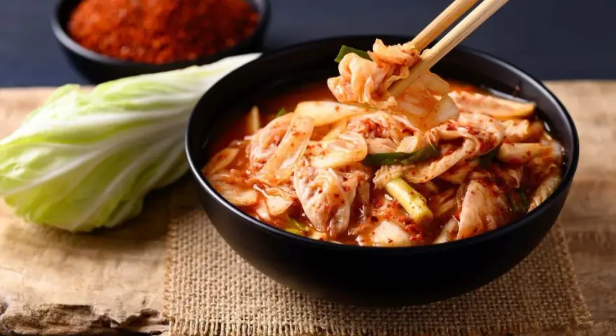 Miseczka z kimchi, pałeczki podnoszące kawałek, kapusta i chili w tle.