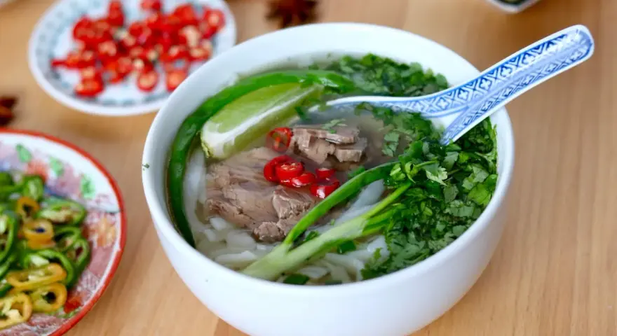 Aromatyczna zupa pho z wołowiną, makaronem ryżowym, świeżą kolendrą, chili i limonką. Obok talerzyki z dodatkami.