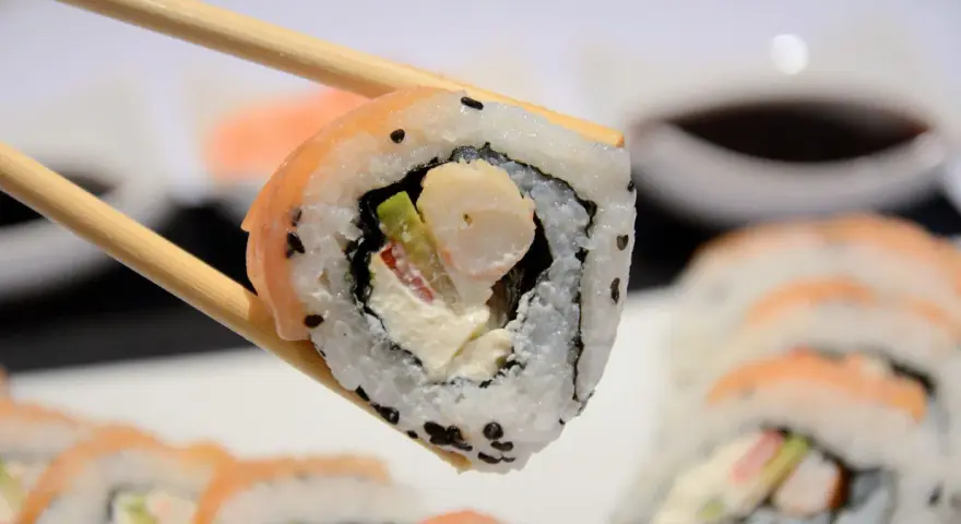 Chopsticks trzymają sushi z krewetkami, awokado i kremowym serkiem. W tle widać więcej roladek sushi.