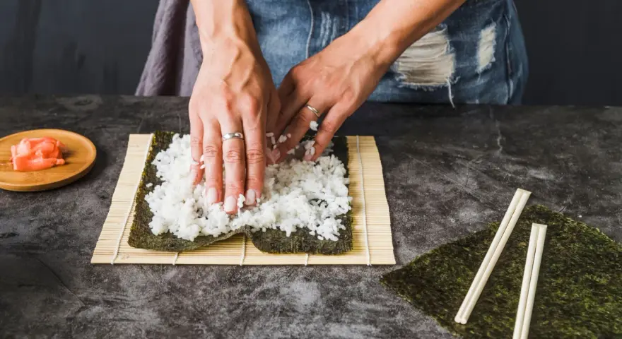 Dłonie rozprowadzają ryż na nori, przygotowując się do zwinięcia sushi. Obok leżą pałeczki i marynowany imbir, idealny dodatek do sushi.
