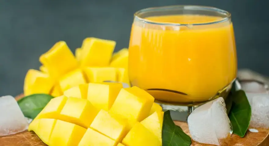 Orzeźwiający mango lassi przepis: pokrojony w kostkę dojrzały mango, szklanka gęstego napoju, kostki lodu i zielone liście.