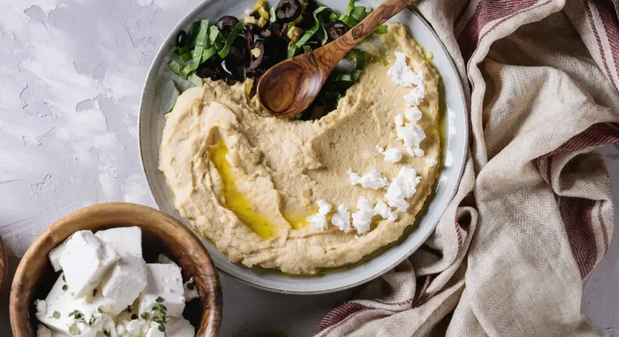 Kremowy hummus z ciecierzycy, posypany fetą i oliwkami, z drewnianą łyżką. Obok miseczka z kostkami sera feta.