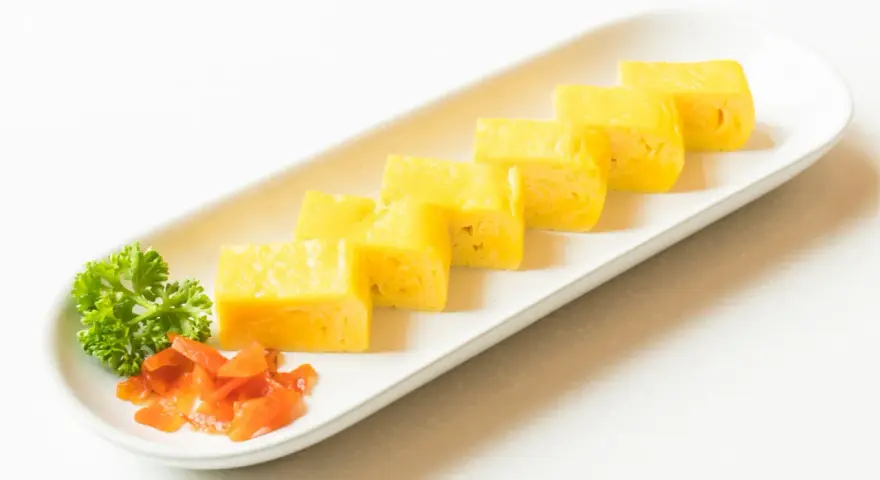 Sześć kawałków tamago, czyli japońskiego omletu, ułożonych na białej desce. Obok natka pietruszki i pomarańczowe dodatki.