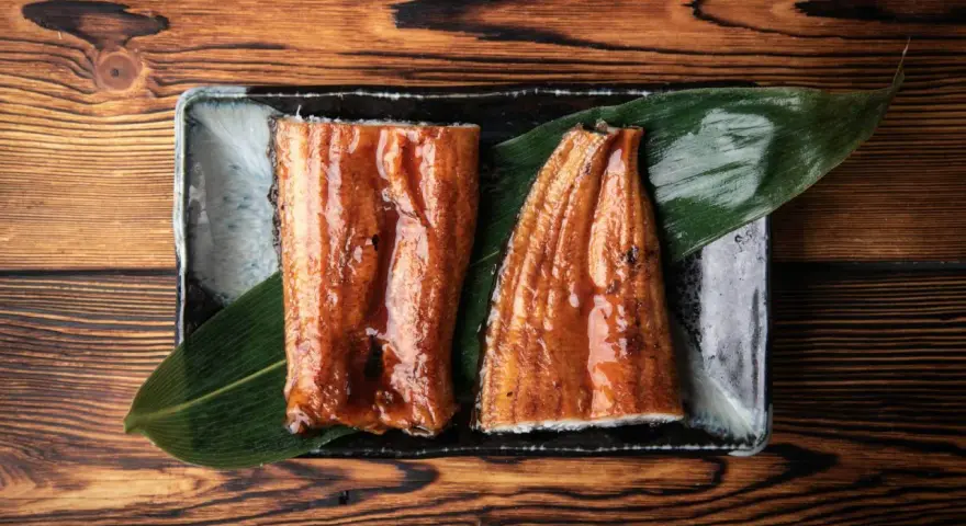 Dwa kawałki błyszczącego, glazurowanego sushi z węgorzem na liściu, podane na czarnej tacce.