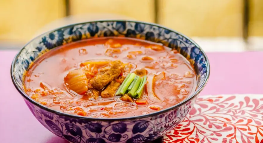 Gorąca zupa z kimchi z kawałkami mięsa i zieloną cebulką, podana w ozdobnej misce.