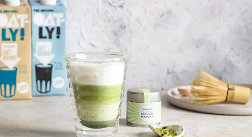 Orzeźwiający iced matcha latte z pianką, obok puszka matcha i bambusowa miotełka. Idealny napój na ciepłe dni.