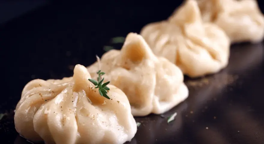 A row of delicate pierożki z krewetkami, garnished with fresh herbs and spices, presented on a dark plate.