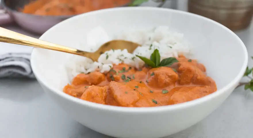 Aksamitny butter chicken masala z ryżem, udekorowany miętą. Złota łyżka zanurzona w daniu.