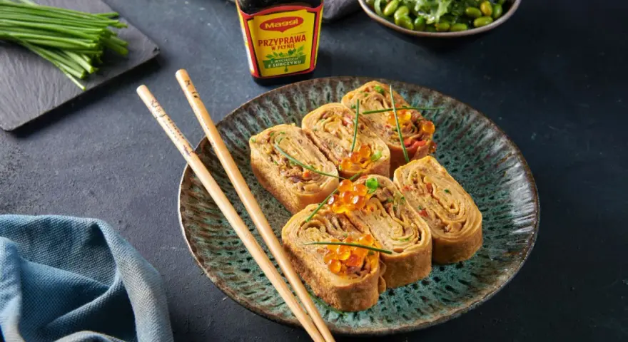 Roladki z omletu do sushi, ozdobione ikrą łososia i szczypiorkiem, obok pałeczki i sosu Maggi.
