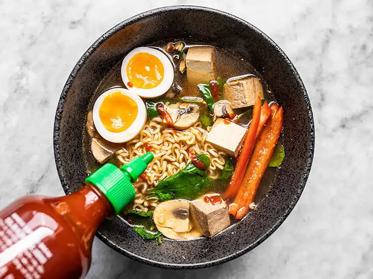 Jak zrobić ramen z zupki chińskiej: miska pełna makaronu, jajka na pół, tofu, grzyby, szpinak i papryka. Sriracha dodaje pikanterii.