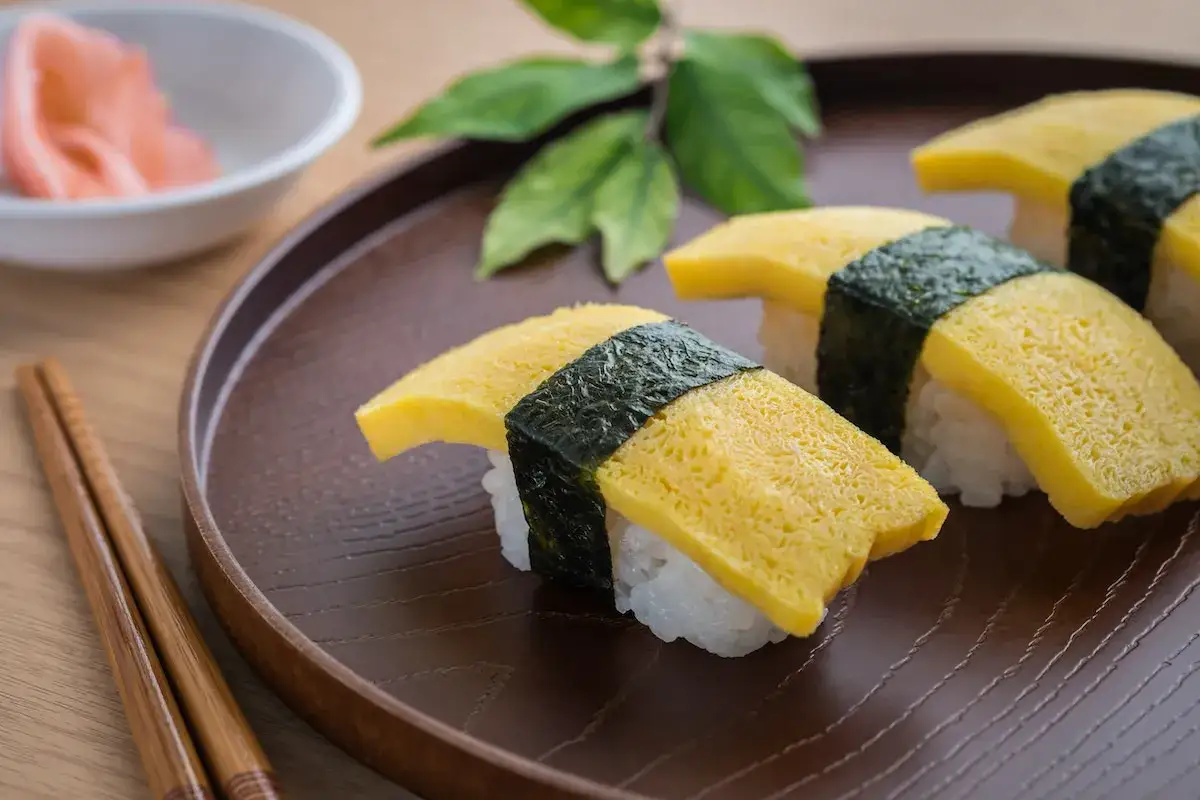 Delikatny omlet do sushi, owinięty w nori, leży na brązowym talerzu obok pałeczek i miseczki z imbirem.