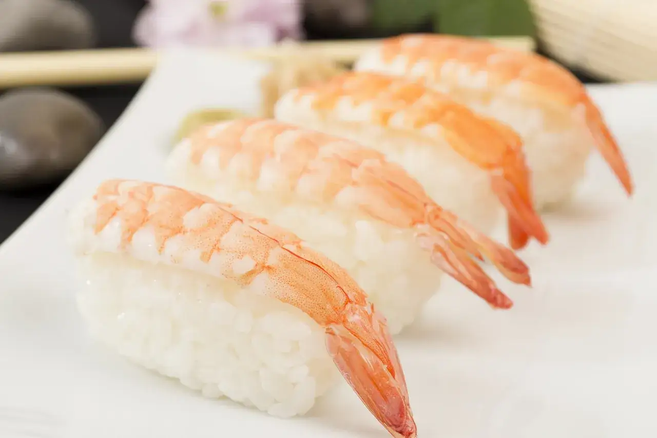 Jak przygotować krewetki do sushi? Oto gotowe nigiri z krewetkami na białym talerzu, z dodatkami w tle.