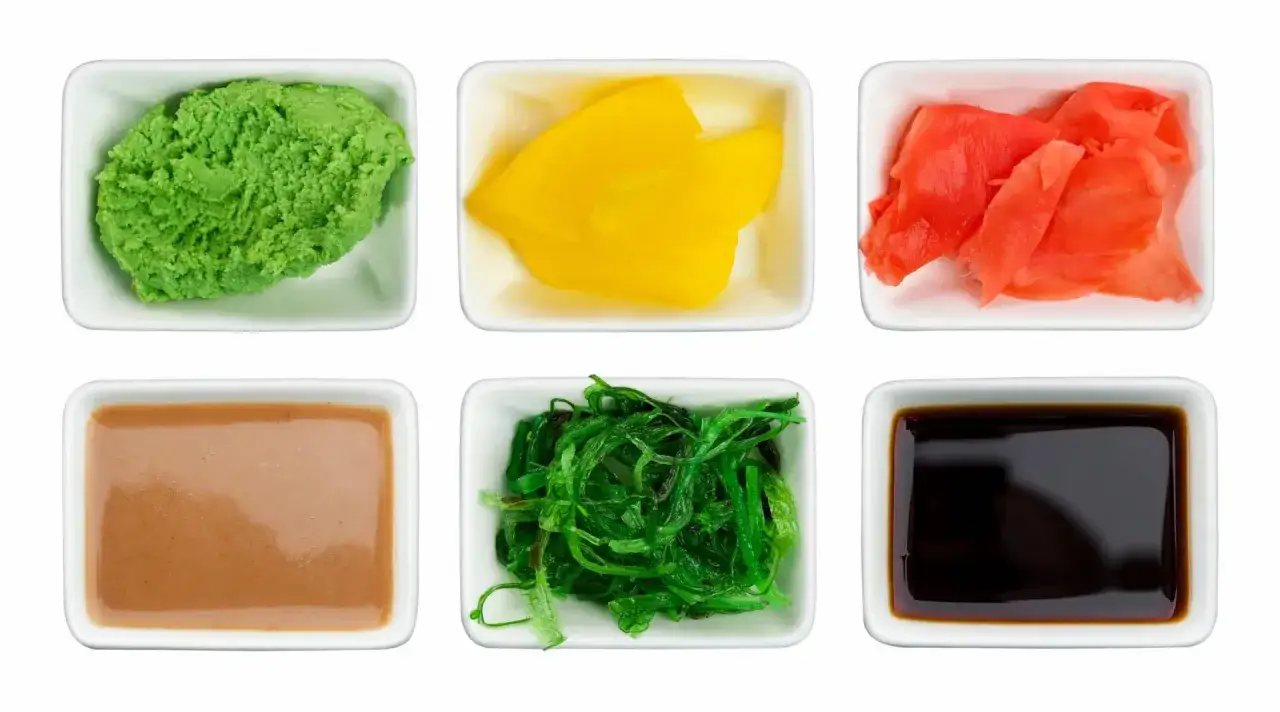 Sześć białych miseczek z dodatkami do sushi: zielone wasabi, żółty imbir marynowany, czerwony imbir marynowany, sos sezamowy, sałatka z wodorostów i sos sojowy. Sos kobe co to?