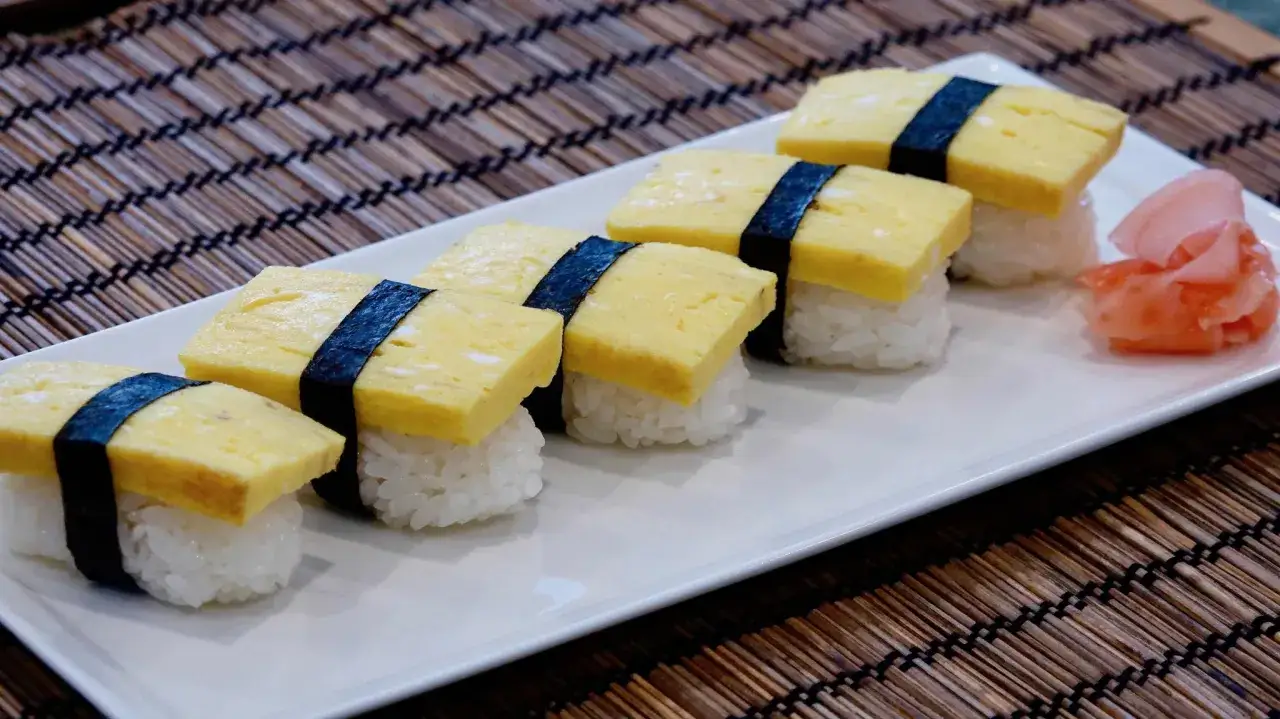 Sześć kawałków sushi z tamago (jajecznica) na ryżu, przewiązane paskiem nori. Obok różowy imbir.