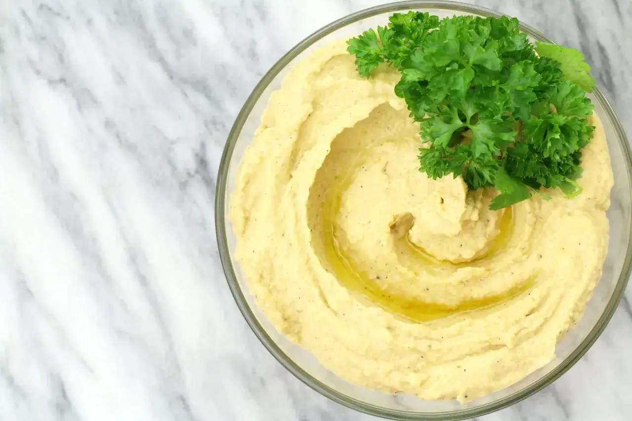Gładki, kremowy hummus z ciecierzycy w szklanej misce, polany oliwą i ozdobiony gałązką natki pietruszki. Idealny do maczania.