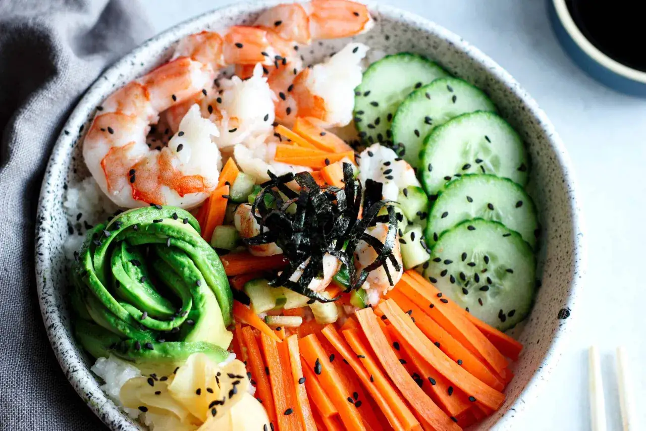 Kolorowa miska z ryżem, awokado, krewetkami, ogórkiem i marchewką, posypana czarnym sezamem. Pyszne sushi z krewetkami.