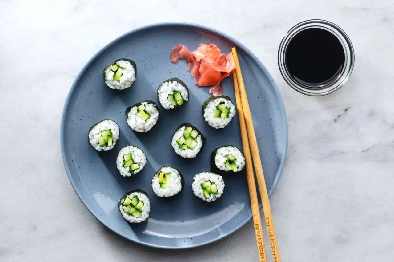 Sushi z ogórkiem, pokazujące, jak pokroić ogórka do sushi. Na talerzu ułożone są rolki sushi, obok pałeczki, marynowany imbir i sos sojowy.