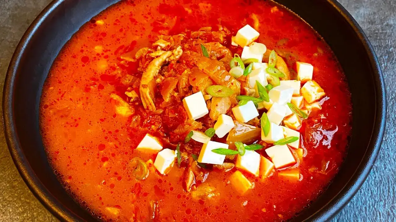 Gorąca, pikantna zupa z kimchi z kostkami tofu i szczypiorkiem, podana w ciemnej misce.