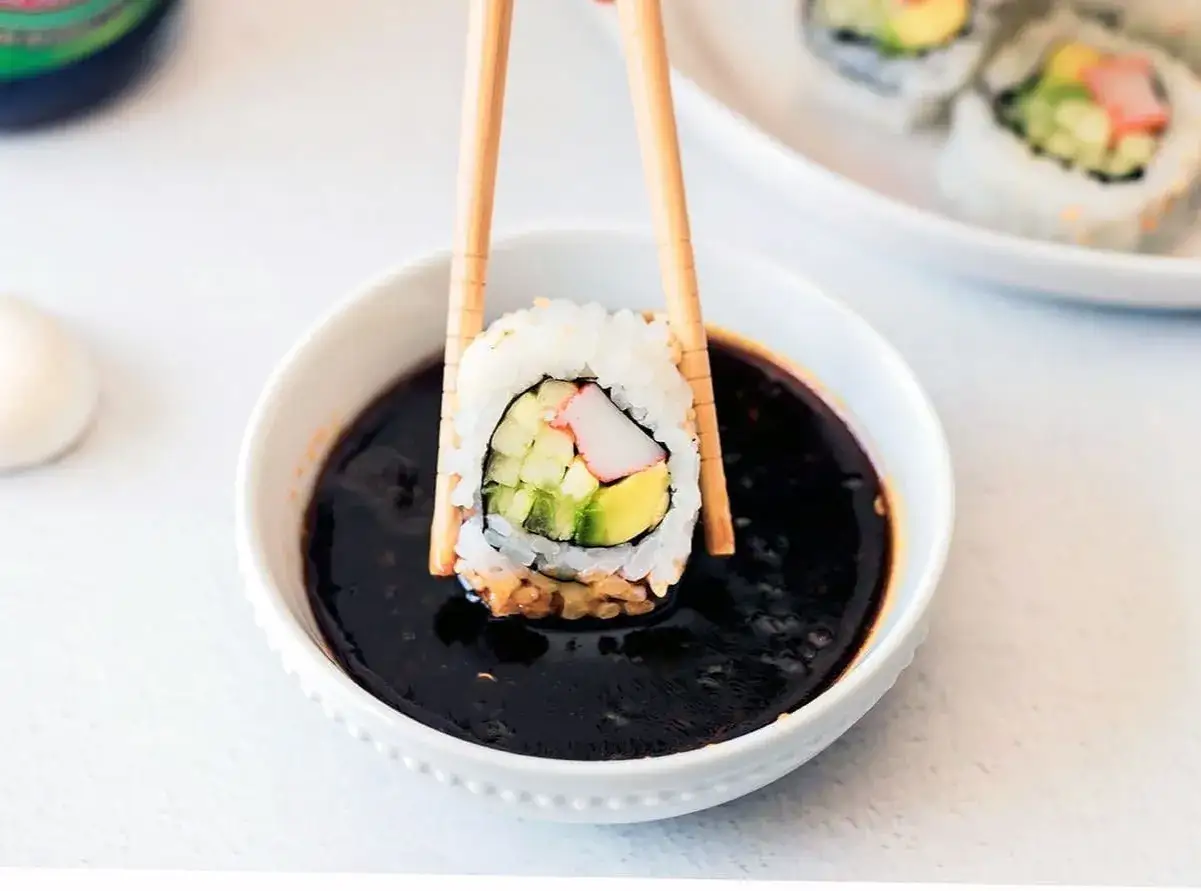 Pałeczkami zanurzam sushi w ciemnym, słodkim sosie. W środku rolki widać awokado, ogórek i paluszek krabowy.