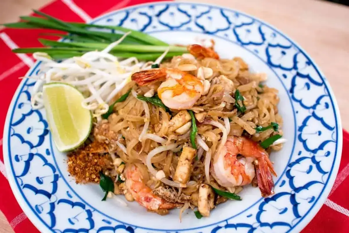 Pyszne danie z makaronem pad thai z krewetkami, kiełkami fasoli mung, szczypiorkiem i ćwiartką limonki na talerzu z niebieskim wzorem.