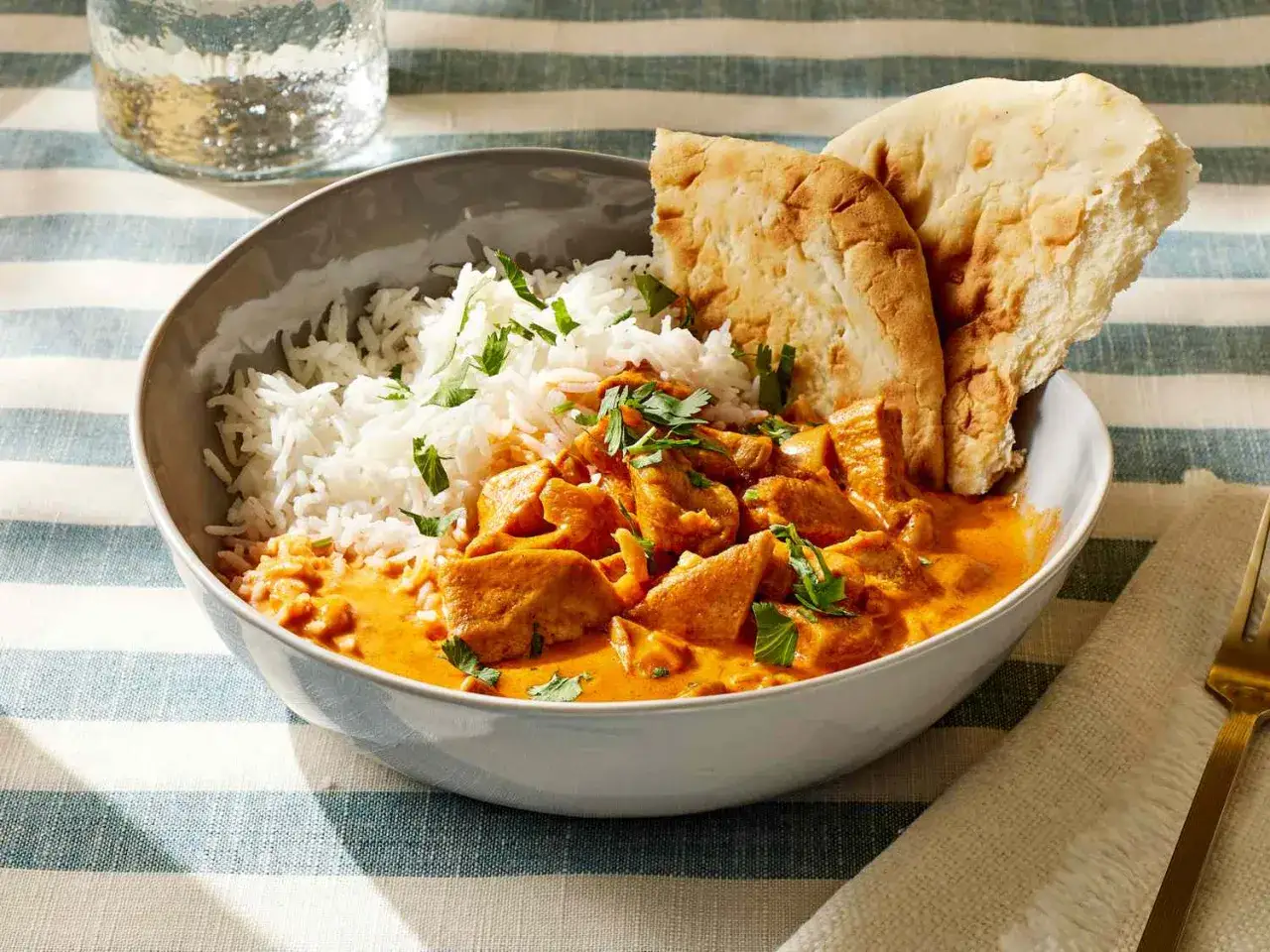 Miseczka z ryżem i kremowym kurczakiem w sosie pomidorowym, posypane natką pietruszki. Obok dwa kawałki chlebka naan. Idealny butter chicken przepis oryginalny.
