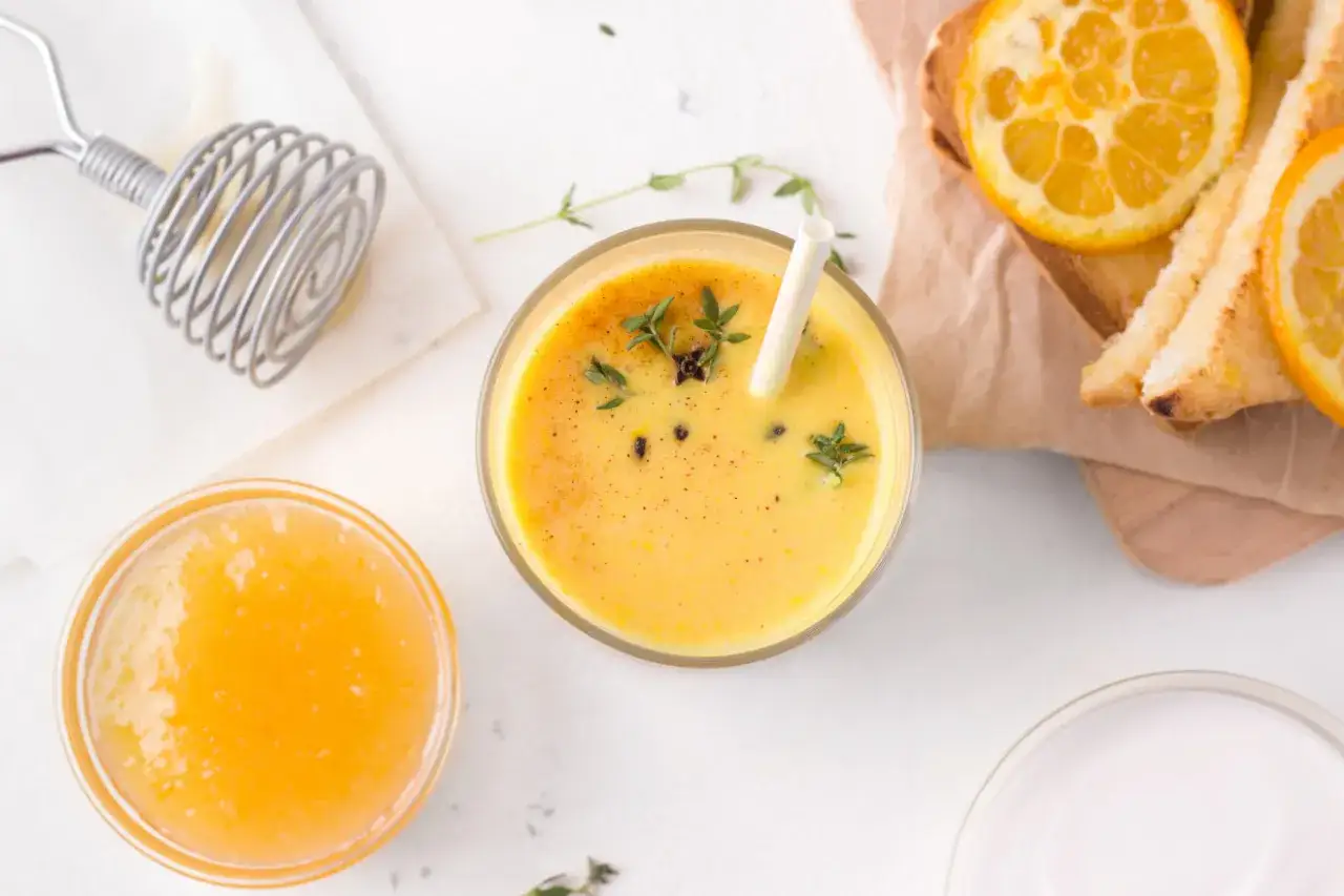 Pyszny mango lassi przepis. Kremowy napój z mango, jogurtem i przyprawami, ozdobiony świeżymi ziołami. Obok miseczka z musem mango i tost z pomarańczą.