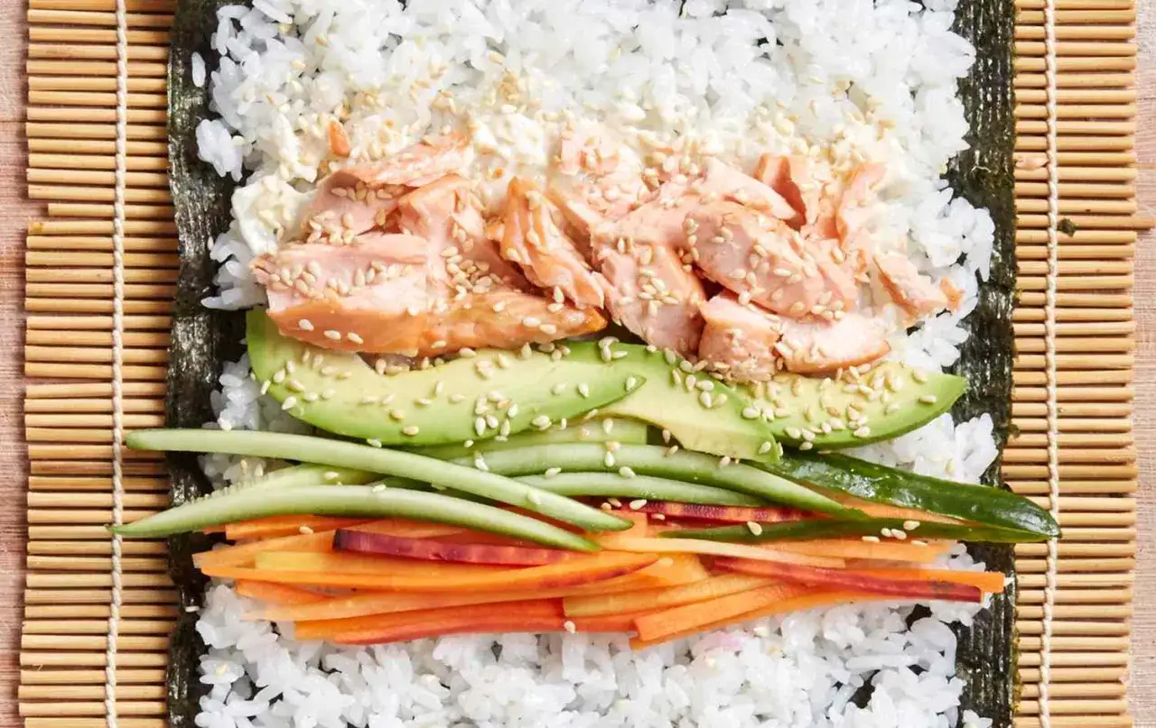 Przygotowanie sushi z pieczonym łososiem: ryż, nori, awokado, ogórek i marchewka czekają na zwinięcie.