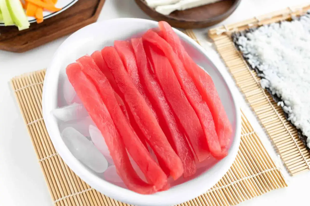 Świeży tuńczyk pokrojony w paski na lodzie, gotowy do przygotowania sushi. Obok mata bambusowa z ryżem.