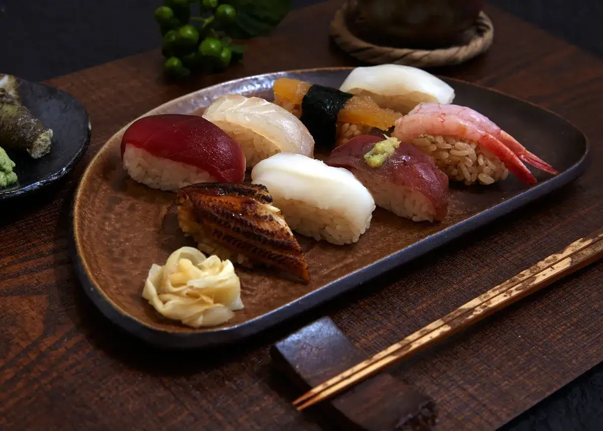 Różnorodne sushi na talerzu: nigiri z tuńczykiem, łososiem, krewetką, kalmarem i węgorzem. Obok marynowany imbir i pałeczki. Tak wygląda sushi.