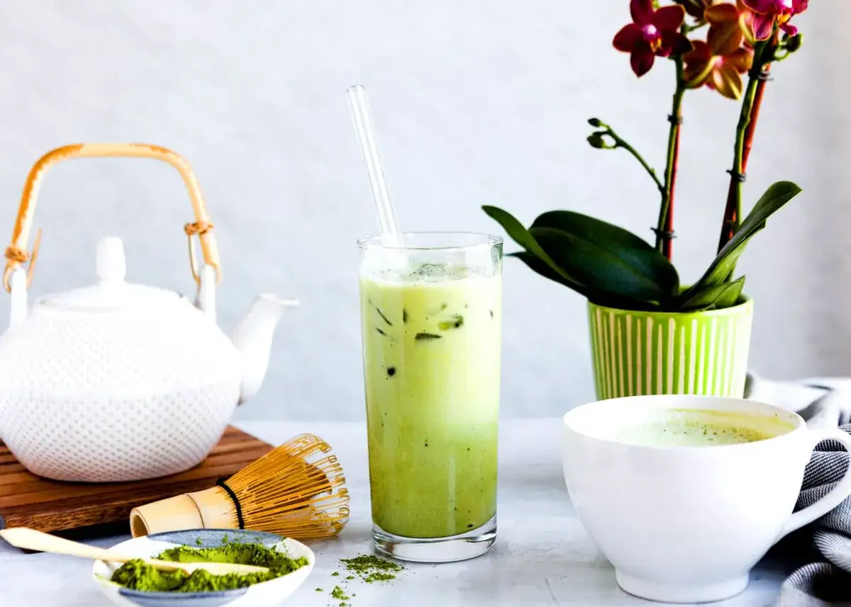 Orzeźwiający iced matcha latte w szklance, obok biały czajniczek, bambusowa miotełka i miseczka z zielonym proszkiem matcha.