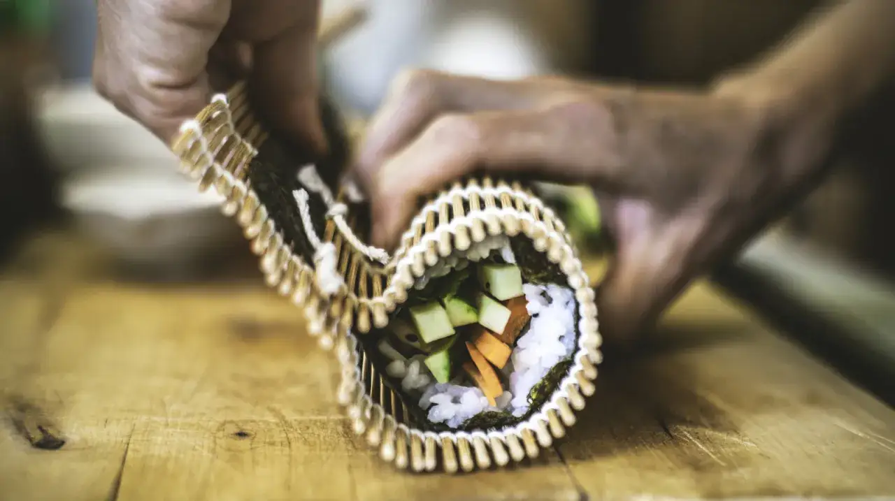 Ręce zwijają sushi z ryżem, ogórkiem i marchewką w wodorostach. Czy sushi ma gluten? Zazwyczaj nie, jeśli nie dodano sosu sojowego z pszenicą.