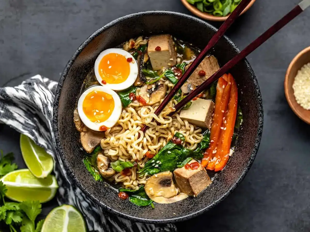 Jak zrobić ramen z zupki chińskiej? Miska pełna makaronu, jajka na pół, tofu, grzyby, szpinak i papryka.