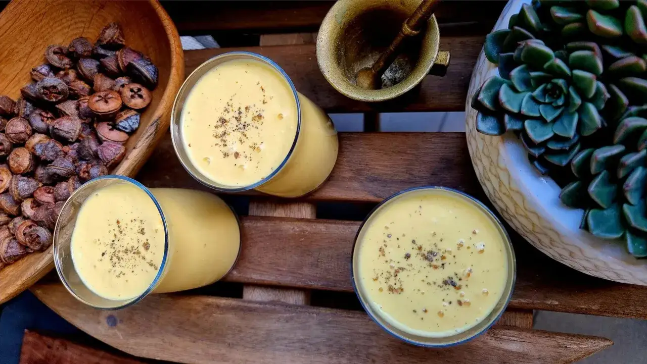 Trzy szklanki kremowego mango lassi, posypane przyprawami. Obok miska z suszonymi owocami, moździerz i sukulent. Idealny mango lassi przepis.