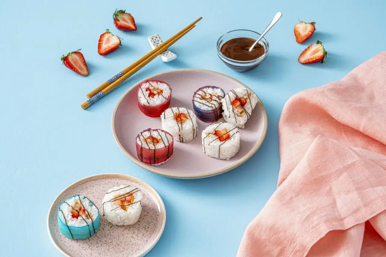 Słodkie sushi z truskawkami i polewą czekoladową, podane na talerzykach z pałeczkami.