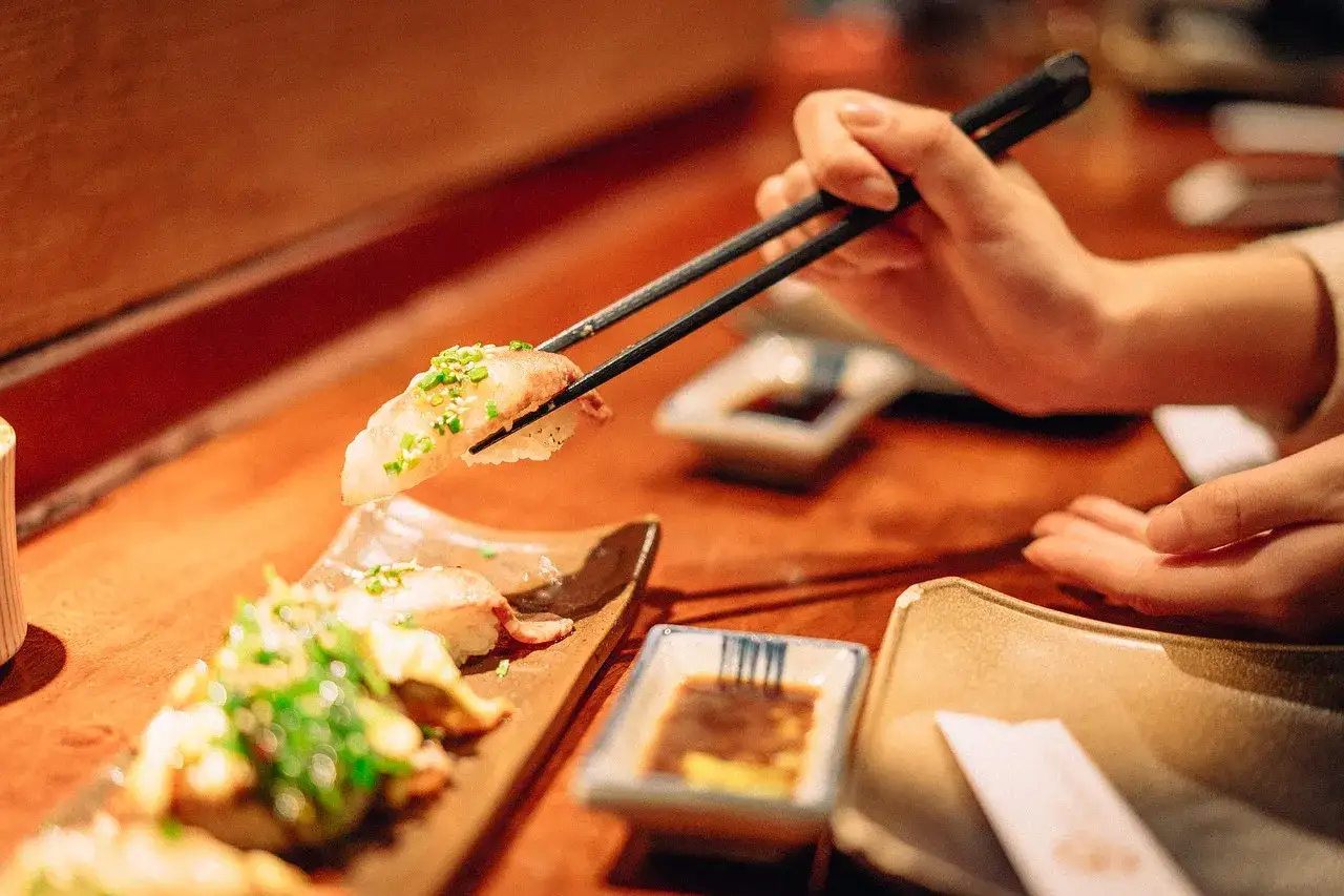 Osoba uczy się, jak jeść pałeczkami, podnosząc kawałek sushi. Na stole widać też inne sushi i sos sojowy.